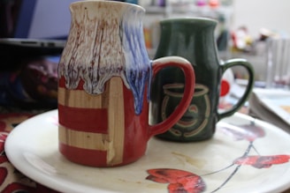 Two mugs are placed on a decorative plate. The mug in the foreground is multicolored with red, beige, and a blue and white dripping glaze at the top. The mug in the background is dark green with a design that resembles a face. They are on a light-colored plate featuring red and black floral patterns, and there is a partially visible laptop in the background.