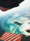 Aerial view of a tropical island surrounded by clear turquoise water. The island features lush greenery and sandy beaches with overwater bungalows extending from the shore. The wing and engine of an aircraft are visible at the top and bottom of the image.