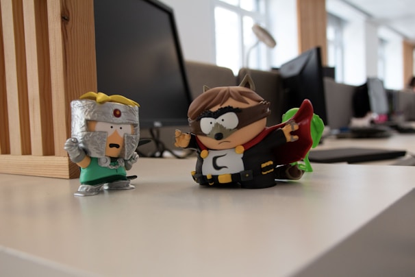 Two cartoonish superhero figurines are placed on a desk in an office setting with computer monitors in the background. One figurine wears a green outfit with a metallic helmet and red gem, while the other wears a dark superhero costume with a mask and cape.