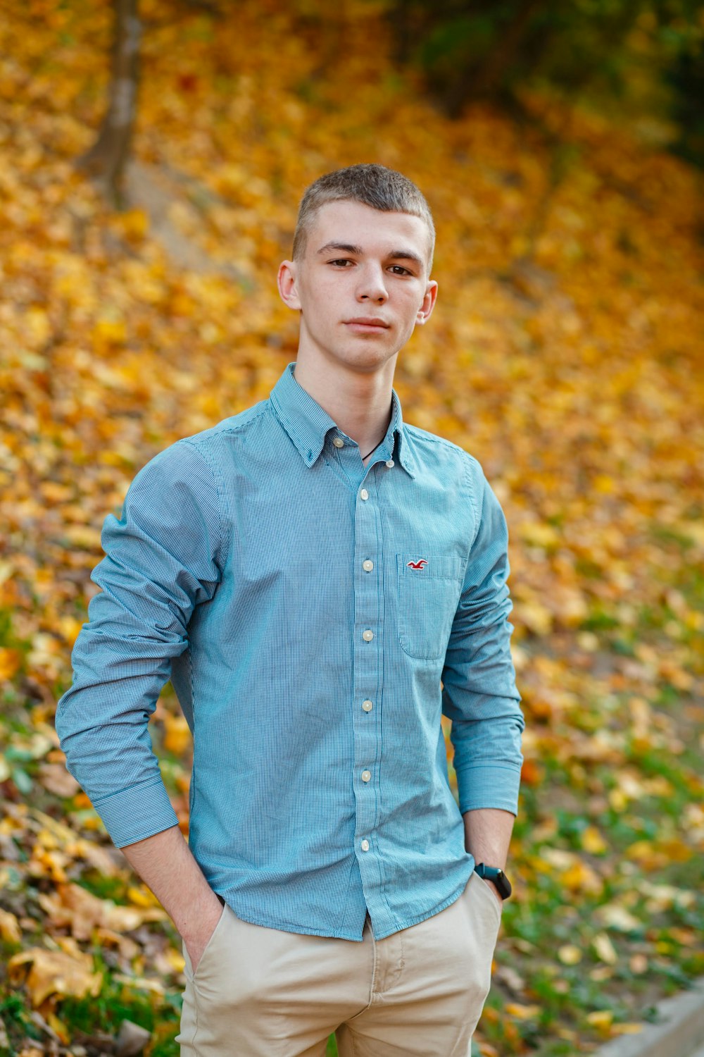 homme en chemise bleue debout près des feuilles brunes pendant la journée