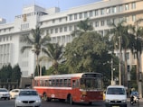 A large, white government building with multiple windows and an emblem on the facade is situated behind a busy road. The front of the building is lined with tall palm trees and green foliage. A red public bus is prominent in the foreground, accompanied by several cars and a motorcyclist.