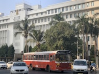A large, white government building with multiple windows and an emblem on the facade is situated behind a busy road. The front of the building is lined with tall palm trees and green foliage. A red public bus is prominent in the foreground, accompanied by several cars and a motorcyclist.