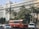 A large, white government building with multiple windows and an emblem on the facade is situated behind a busy road. The front of the building is lined with tall palm trees and green foliage. A red public bus is prominent in the foreground, accompanied by several cars and a motorcyclist.