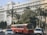 A large, white government building with multiple windows and an emblem on the facade is situated behind a busy road. The front of the building is lined with tall palm trees and green foliage. A red public bus is prominent in the foreground, accompanied by several cars and a motorcyclist.
