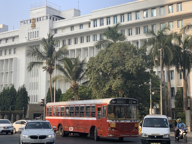 A large, white government building with multiple windows and an emblem on the facade is situated behind a busy road. The front of the building is lined with tall palm trees and green foliage. A red public bus is prominent in the foreground, accompanied by several cars and a motorcyclist.