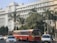 A large, white government building with multiple windows and an emblem on the facade is situated behind a busy road. The front of the building is lined with tall palm trees and green foliage. A red public bus is prominent in the foreground, accompanied by several cars and a motorcyclist.