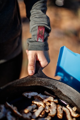 A person is holding a cast-iron skillet filled with sliced mushrooms, with a focus on the hand gripping the skillet's handle. The individual is wearing a long-sleeved garment with a decorative emblem near the wrist. A bright blue container is visible in the background, hinting at an outdoor setting.