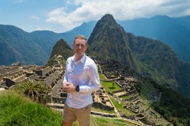 A person stands in the foreground wearing a white shirt and khaki shorts, smiling against a backdrop of the ancient ruins of Machu Picchu, with its terraces, green mountain landscape, and dramatic peaks under a blue sky.