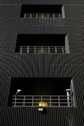 A dark building facade with vertical corrugated siding, featuring multiple levels with recessed openings. Each opening is guarded by metal railings, and two security cameras flank the bottommost level. A warm yellow light emanates from behind the lower railing, creating a stark contrast with the dark exterior.