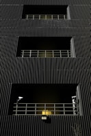 A dark building facade with vertical corrugated siding, featuring multiple levels with recessed openings. Each opening is guarded by metal railings, and two security cameras flank the bottommost level. A warm yellow light emanates from behind the lower railing, creating a stark contrast with the dark exterior.