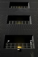 A dark building facade with vertical corrugated siding, featuring multiple levels with recessed openings. Each opening is guarded by metal railings, and two security cameras flank the bottommost level. A warm yellow light emanates from behind the lower railing, creating a stark contrast with the dark exterior.