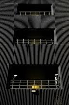 A dark building facade with vertical corrugated siding, featuring multiple levels with recessed openings. Each opening is guarded by metal railings, and two security cameras flank the bottommost level. A warm yellow light emanates from behind the lower railing, creating a stark contrast with the dark exterior.