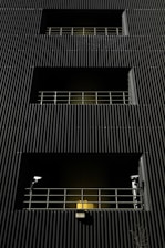 A dark building facade with vertical corrugated siding, featuring multiple levels with recessed openings. Each opening is guarded by metal railings, and two security cameras flank the bottommost level. A warm yellow light emanates from behind the lower railing, creating a stark contrast with the dark exterior.