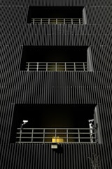 A dark building facade with vertical corrugated siding, featuring multiple levels with recessed openings. Each opening is guarded by metal railings, and two security cameras flank the bottommost level. A warm yellow light emanates from behind the lower railing, creating a stark contrast with the dark exterior.
