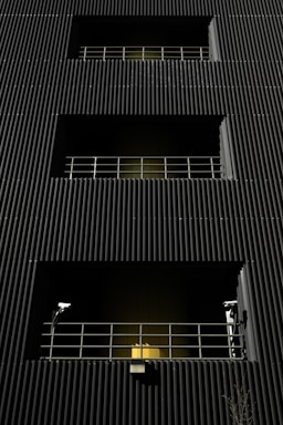 A dark building facade with vertical corrugated siding, featuring multiple levels with recessed openings. Each opening is guarded by metal railings, and two security cameras flank the bottommost level. A warm yellow light emanates from behind the lower railing, creating a stark contrast with the dark exterior.