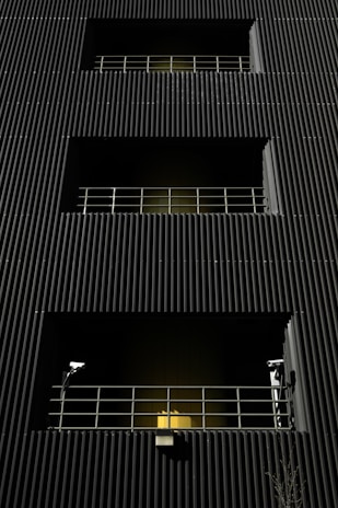 A dark building facade with vertical corrugated siding, featuring multiple levels with recessed openings. Each opening is guarded by metal railings, and two security cameras flank the bottommost level. A warm yellow light emanates from behind the lower railing, creating a stark contrast with the dark exterior.