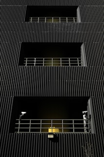 A dark building facade with vertical corrugated siding, featuring multiple levels with recessed openings. Each opening is guarded by metal railings, and two security cameras flank the bottommost level. A warm yellow light emanates from behind the lower railing, creating a stark contrast with the dark exterior.