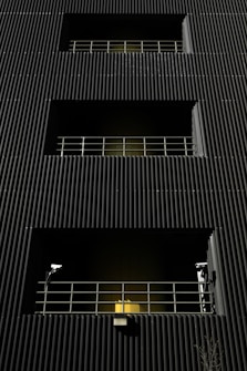 A dark building facade with vertical corrugated siding, featuring multiple levels with recessed openings. Each opening is guarded by metal railings, and two security cameras flank the bottommost level. A warm yellow light emanates from behind the lower railing, creating a stark contrast with the dark exterior.