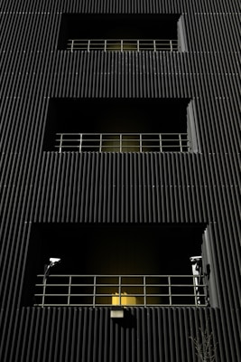 A dark building facade with vertical corrugated siding, featuring multiple levels with recessed openings. Each opening is guarded by metal railings, and two security cameras flank the bottommost level. A warm yellow light emanates from behind the lower railing, creating a stark contrast with the dark exterior.