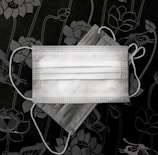 Two surgical face masks lie on a dark floral-patterned background. The masks are white and overlapping, with visible ear loops. The design features lotus-like flowers in shades of gray.