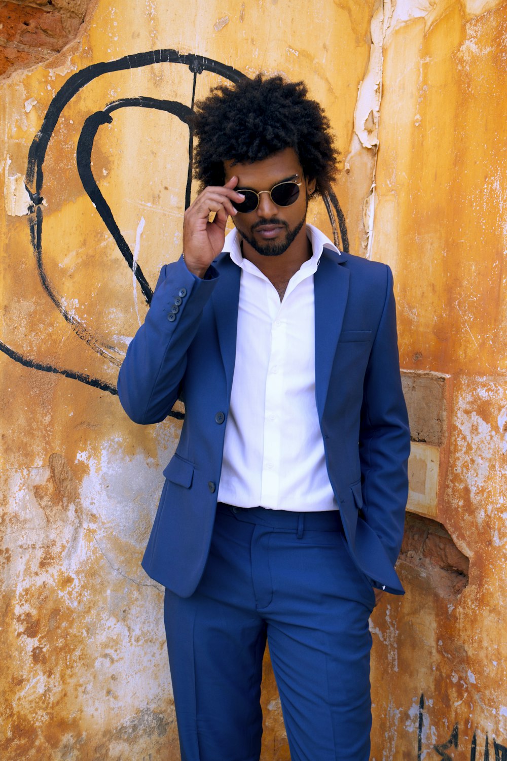 파란색 양복 재킷을 입고 갈색 콘크리트 벽 옆에 서 있는 검은 선글라스를 쓴 남자