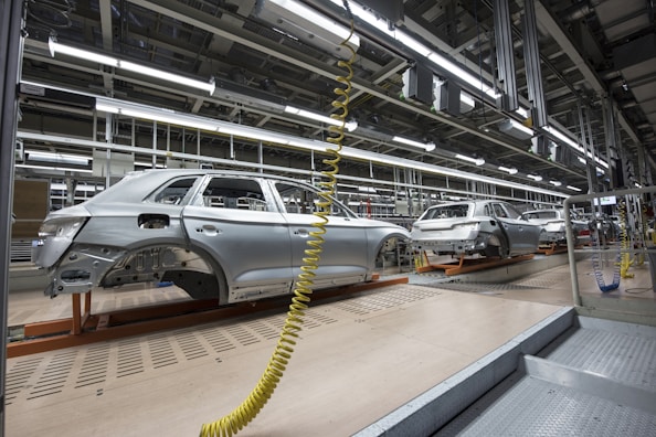 An assembly line in a car manufacturing plant with several partially assembled vehicle frames lined up. Robotic equipment and wiring are visible, emphasizing the mechanical aspect of the production process. The environment is clean and organized, with bright overhead lighting illuminating the industrial setting.