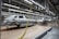 An assembly line in a car manufacturing plant with several partially assembled vehicle frames lined up. Robotic equipment and wiring are visible, emphasizing the mechanical aspect of the production process. The environment is clean and organized, with bright overhead lighting illuminating the industrial setting.