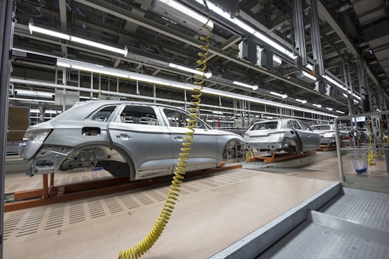 An assembly line in a car manufacturing plant with several partially assembled vehicle frames lined up. Robotic equipment and wiring are visible, emphasizing the mechanical aspect of the production process. The environment is clean and organized, with bright overhead lighting illuminating the industrial setting.