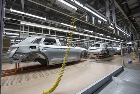 An assembly line in a car manufacturing plant with several partially assembled vehicle frames lined up. Robotic equipment and wiring are visible, emphasizing the mechanical aspect of the production process. The environment is clean and organized, with bright overhead lighting illuminating the industrial setting.