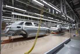 An assembly line in a car manufacturing plant with several partially assembled vehicle frames lined up. Robotic equipment and wiring are visible, emphasizing the mechanical aspect of the production process. The environment is clean and organized, with bright overhead lighting illuminating the industrial setting.
