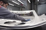 A person is polishing or inspecting a metallic car part in a factory setting. The environment looks industrial with metal structures, overhead lights, and multiple stations. The car part being handled appears to be a large metal panel, possibly a door or hood of a vehicle.