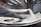 A person is polishing or inspecting a metallic car part in a factory setting. The environment looks industrial with metal structures, overhead lights, and multiple stations. The car part being handled appears to be a large metal panel, possibly a door or hood of a vehicle.