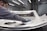 A person is polishing or inspecting a metallic car part in a factory setting. The environment looks industrial with metal structures, overhead lights, and multiple stations. The car part being handled appears to be a large metal panel, possibly a door or hood of a vehicle.