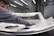 A person is polishing or inspecting a metallic car part in a factory setting. The environment looks industrial with metal structures, overhead lights, and multiple stations. The car part being handled appears to be a large metal panel, possibly a door or hood of a vehicle.
