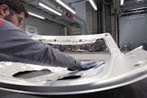 A person is polishing or inspecting a metallic car part in a factory setting. The environment looks industrial with metal structures, overhead lights, and multiple stations. The car part being handled appears to be a large metal panel, possibly a door or hood of a vehicle.