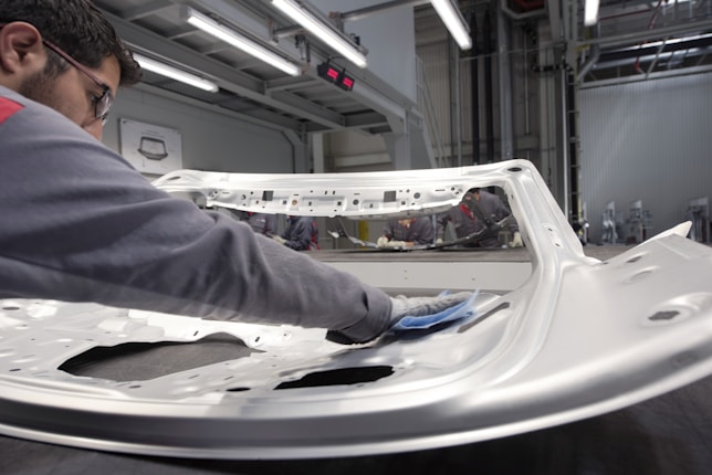 A person is polishing or inspecting a metallic car part in a factory setting. The environment looks industrial with metal structures, overhead lights, and multiple stations. The car part being handled appears to be a large metal panel, possibly a door or hood of a vehicle.