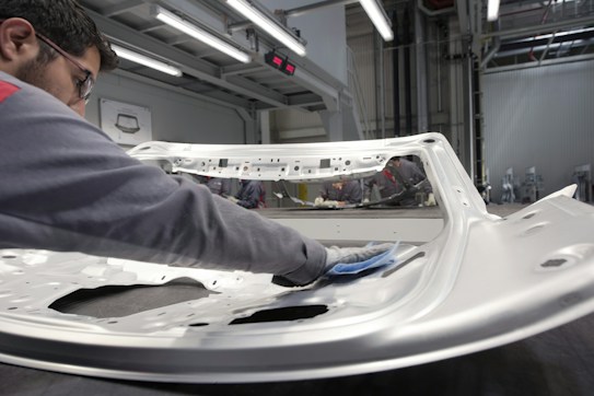 A person is polishing or inspecting a metallic car part in a factory setting. The environment looks industrial with metal structures, overhead lights, and multiple stations. The car part being handled appears to be a large metal panel, possibly a door or hood of a vehicle.
