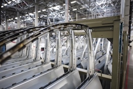Multiple car doors and frames are lined up in a row inside an industrial facility with a high ceiling featuring metal beams and structures.