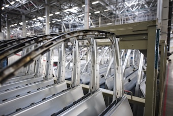 Multiple car doors and frames are lined up in a row inside an industrial facility with a high ceiling featuring metal beams and structures.