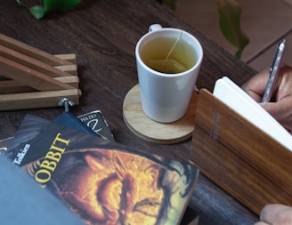 A white cup of tea with a tea bag on a wooden coaster sits next to a hand writing in a notebook. Nearby, there is a stack of books, including a prominent copy of 'The Hobbit' with a vivid cover featuring a dragon. The setting is a wooden table, and wooden clamps or stands are visible in the background.