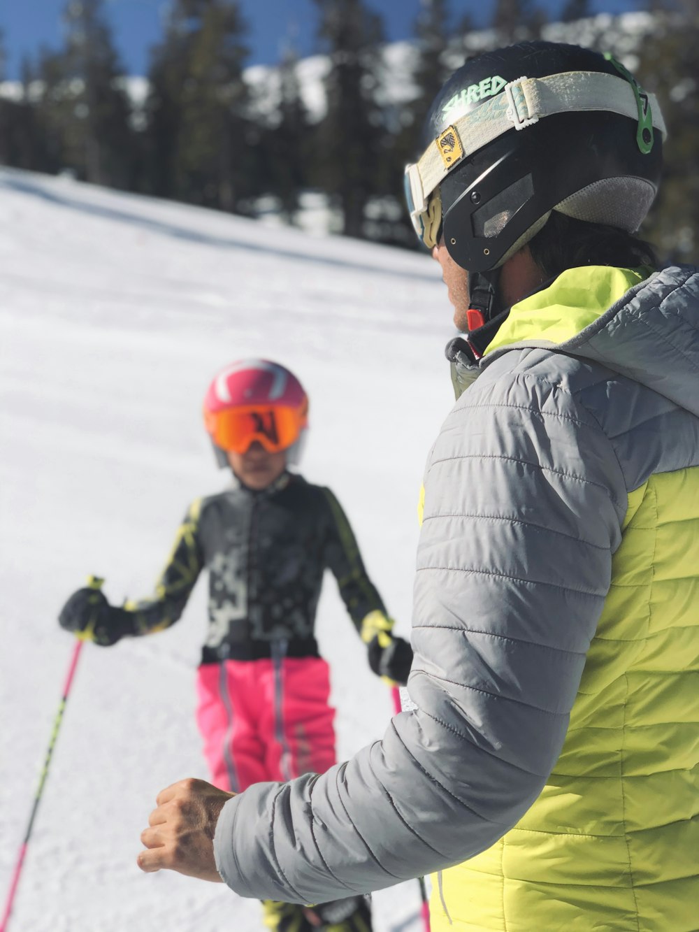 Man met gele jas die zijn kind met roze skiuitrusting opwacht
