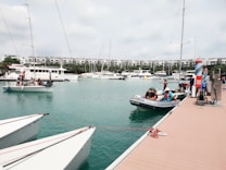 A marina with several boats docked on a calm, turquoise water surface. People are seen on a small boat near the dock and others are preparing to board or disembark. In the background, there is a row of modern residential buildings with lush greenery.