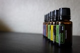 Several amber glass bottles with black caps are aligned in a row on a dark surface. Each bottle has a label indicating the type of essential oil it contains, such as Tea Tree and Lemon. The background is blurred, creating a shallow depth of field.
