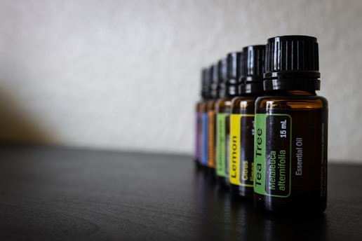 Several amber glass bottles with black caps are aligned in a row on a dark surface. Each bottle has a label indicating the type of essential oil it contains, such as Tea Tree and Lemon. The background is blurred, creating a shallow depth of field.