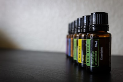 Several amber glass bottles with black caps are aligned in a row on a dark surface. Each bottle has a label indicating the type of essential oil it contains, such as Tea Tree and Lemon. The background is blurred, creating a shallow depth of field.