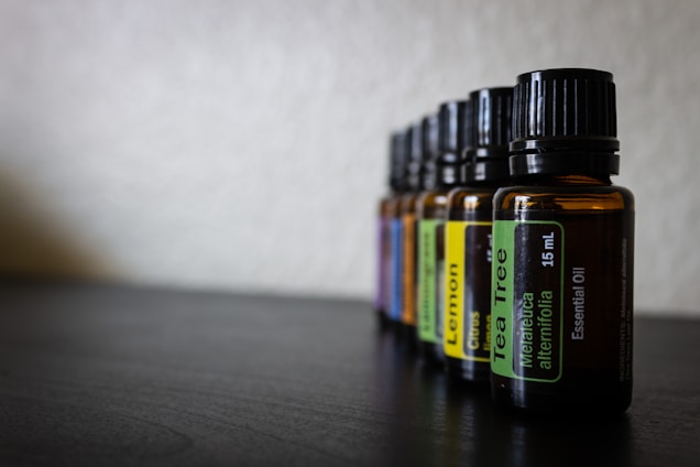 Several amber glass bottles with black caps are aligned in a row on a dark surface. Each bottle has a label indicating the type of essential oil it contains, such as Tea Tree and Lemon. The background is blurred, creating a shallow depth of field.