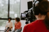 A professional camera operator wearing headphones is filming two people who are sitting in a well-lit room with large windows. The camera display shows the two individuals slightly blurred in the background.