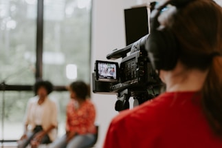 A professional camera operator wearing headphones is filming two people who are sitting in a well-lit room with large windows. The camera display shows the two individuals slightly blurred in the background.