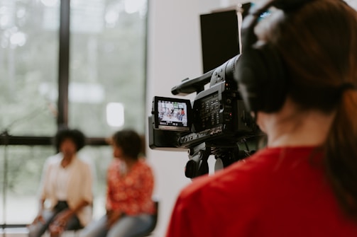 A professional camera operator wearing headphones is filming two people who are sitting in a well-lit room with large windows. The camera display shows the two individuals slightly blurred in the background.