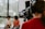 A professional camera operator wearing headphones is filming two people who are sitting in a well-lit room with large windows. The camera display shows the two individuals slightly blurred in the background.